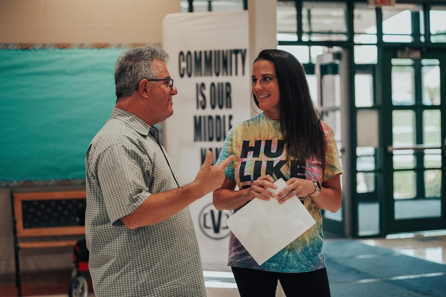 Two people in conversation near the entrance doors, beside a banner reading Community Is Our Middle Name